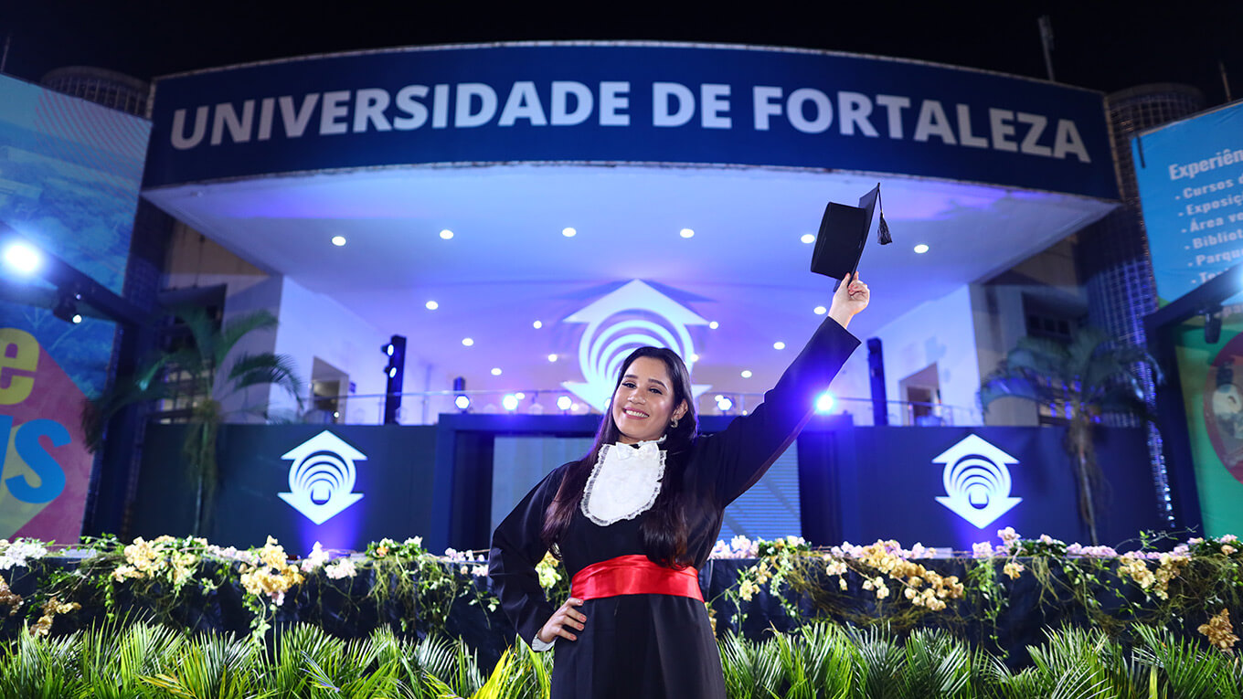 Jovem ergue chapéu de formatura e sorri em frente ao Centro de Convivência