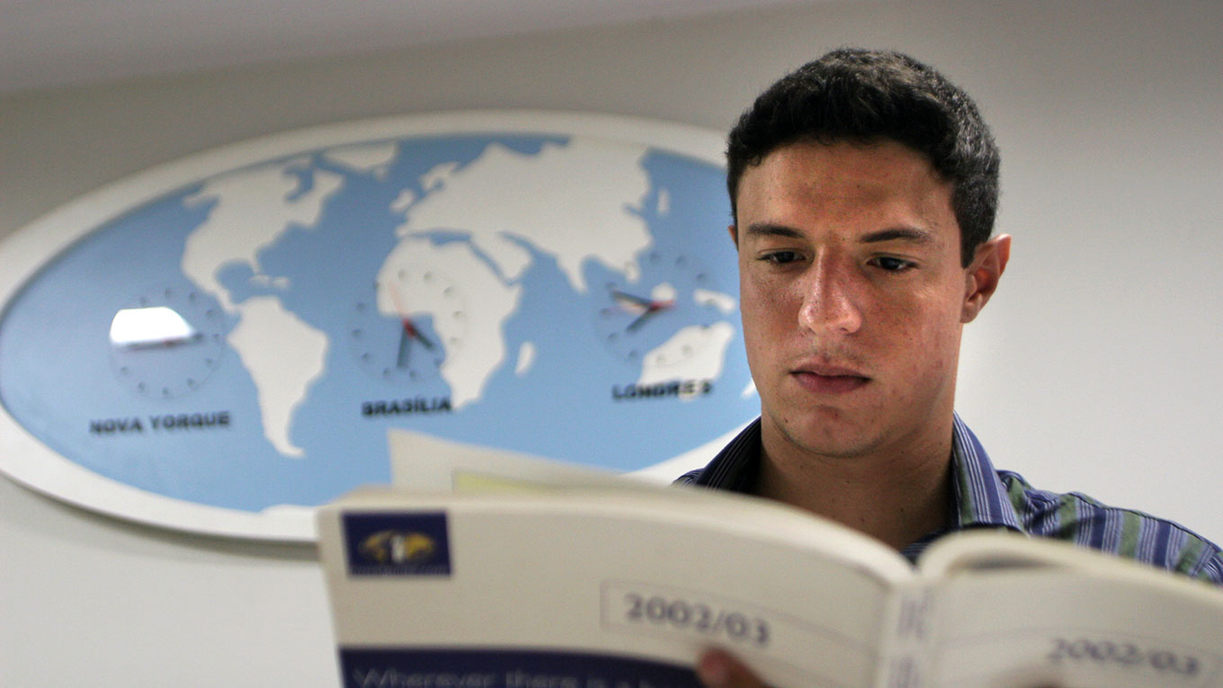 aluno pesquisando em livro com relógio atrás informando fusos horários de outros países do mundo