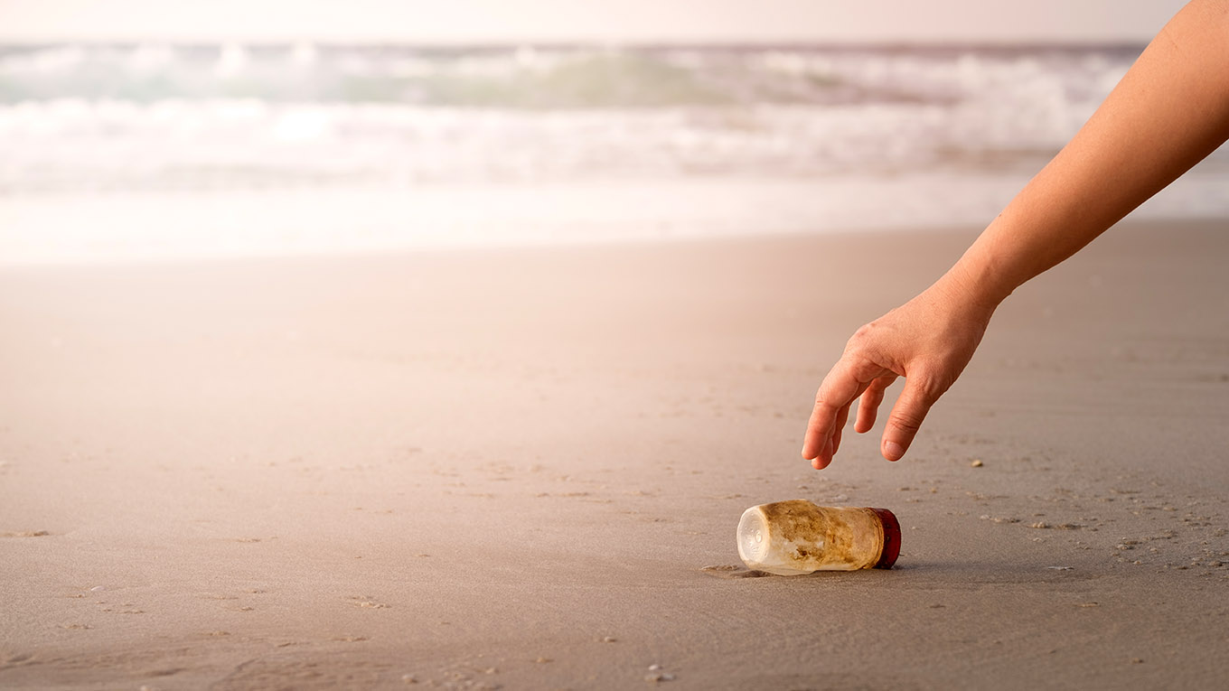 mão de uma pessoa recolhendo uma garrafa plástica suja na beira da praia