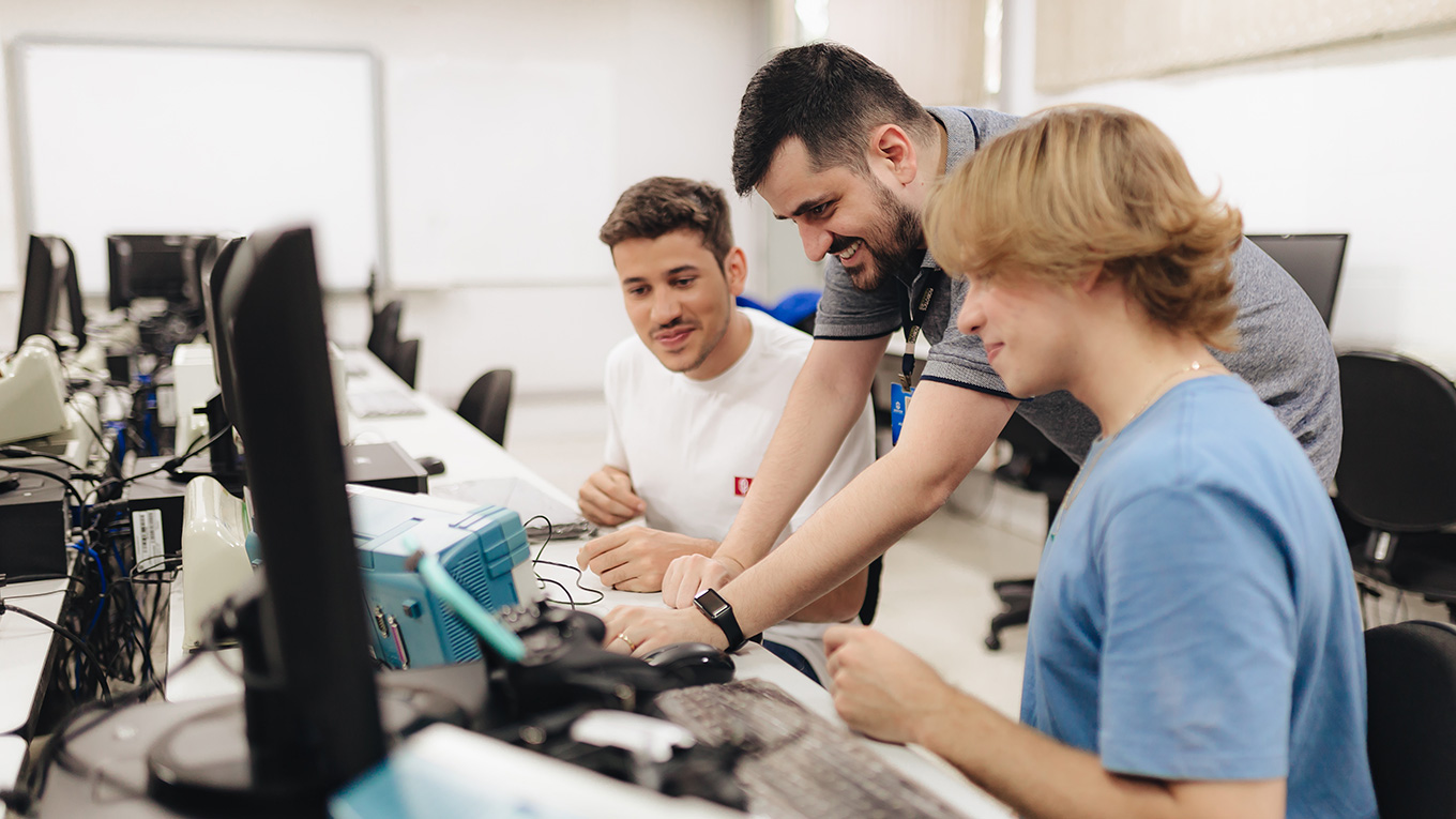 Professor ensina alunos enquanto manipula equipamento em meio a computadores