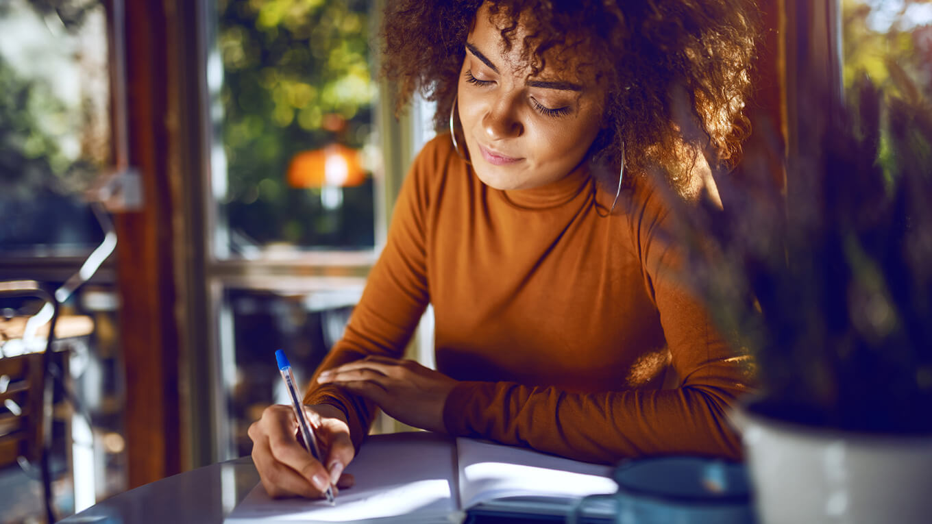 Uma mulher em destaque sorrindo e ela está escrevendo em um caderno.