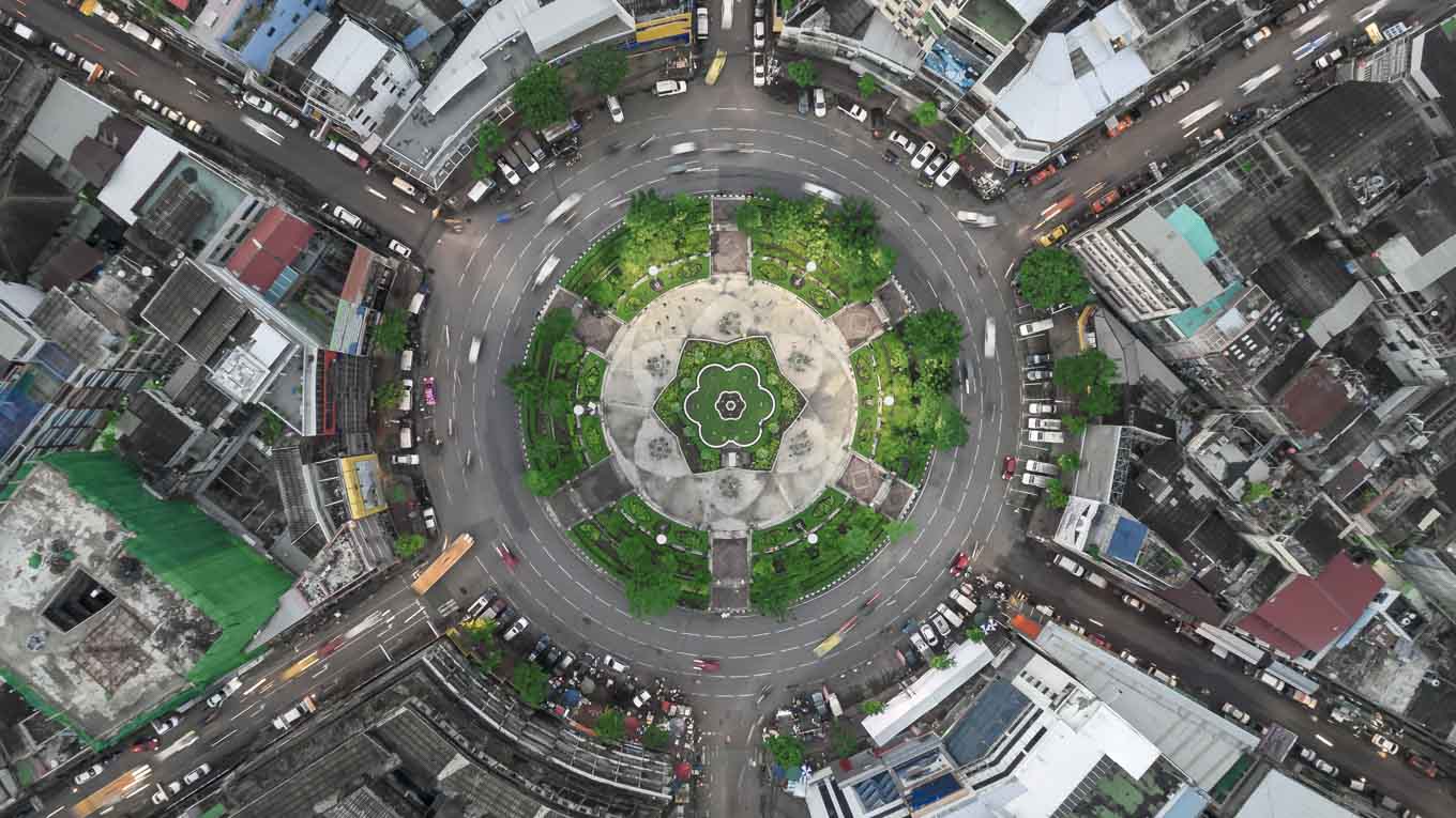 Uma rotatória arborizada, no centro de uma cidade, é vista de cima.