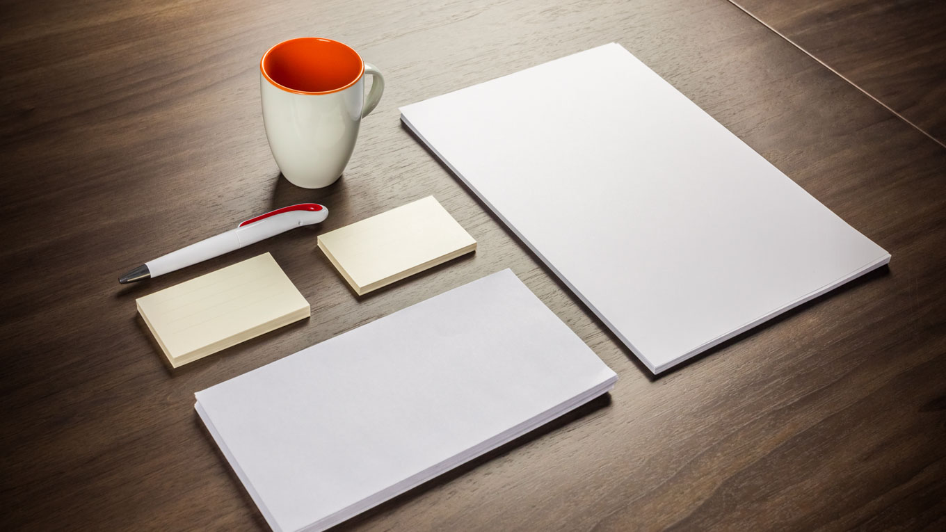 Sobre uma mesa estão dispostos papéis em branco, caneta e caneca.